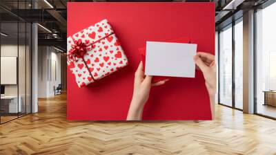 First person top view photo of st valentine's day decor girl's hands holding envelope and card over giftbox in white wrapping paper heart pattern red star bow on isolated red background with copyspace Wall mural