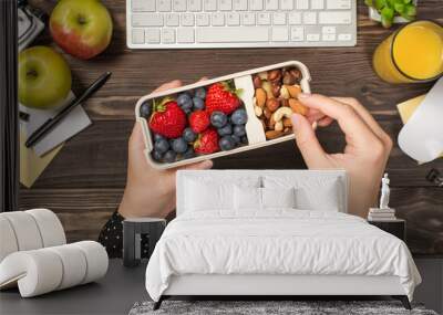 First person top view photo of woman's hands holding lunchbox with healthy meal nuts and berries over apples glass of juice flowerpot stationery keyboard mouse on isolated dark wooden table background Wall mural