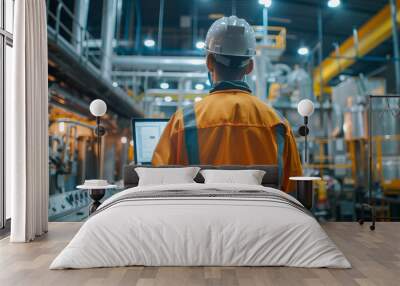 person from behind, wearing a safety helmet and an orange high-visibility jacket, holding laptop, standing in an industrial setting with large metallic structures and machinery in a plant environment Wall mural