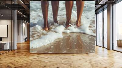 Rear back view of two women, female legs and barefoot feet standing on sand beach, sea or ocean water waves with foam splash Wall mural