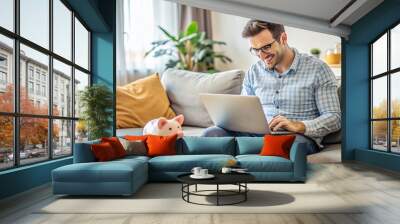 Young adult sitting on couch with laptop open to online banking website, surrounded by financial papers and piggy bank, planning financial future. Wall mural