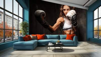 Sport young woman wearing boxing gloves posing in combat stance looking at camera. Fit young female boxer ready for fight on studio dark background. Copy space Wall mural