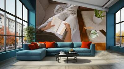 Man in cozy home wrapped in blanked relaxing on sofa holding book and reading. Warm sunlight entering living room. Wall mural