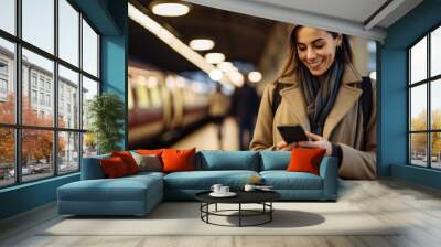 Smiling attractive woman looking at his smart phone at a train station. Wall mural