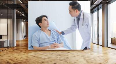 Senior doctor and young male patient who lie on the bed while checking pulse, consult and explain with nurse taking note and supporting in hospital wards. Wall mural