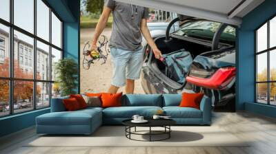 Handsome man getting ready for a vacations, loading his backpack in a car trunk, together with a drone and other camera equipment. Wall mural