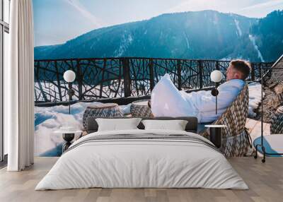 Young man in a robe chilling on a open terrace in the SPA with an amazing mountains view during winter time. Ski resort. Wall mural