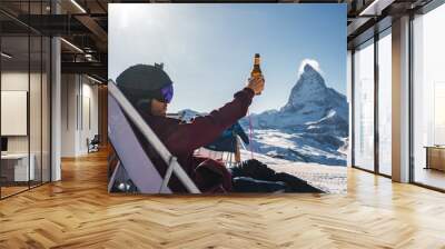 Young snowboarder cheering with a beer after skiing day in a bar or a cafe at the Zermatt ski resort in Swiss Alps with a fabulous view of the Matterhorn. Winter vacation concept. Wall mural