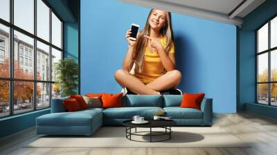 Cheerful teenager girl listening to music with headphones and cellphone on blue background Wall mural