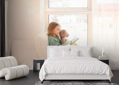 Cute little girl reading book while sitting on window sill at home Wall mural