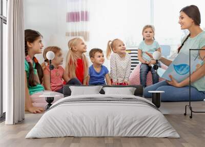 Female teacher reading book to little children in kindergarten Wall mural