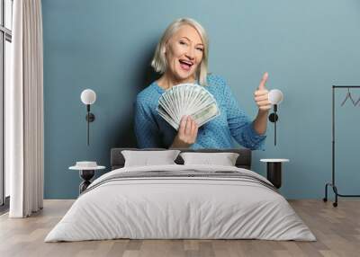 Happy mature woman with money showing thumb-up gesture on color background Wall mural