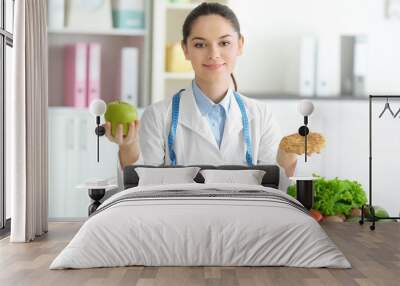 Young female nutritionist holding tasty cookie and apple while sitting at table in her office Wall mural