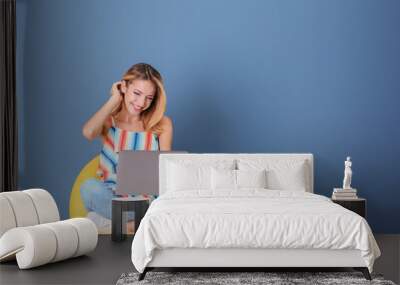 Young woman with laptop in armchair against blue background Wall mural