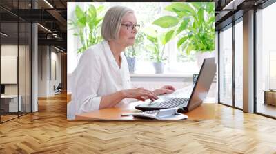 Senior woman at home with a calculator and lots of receipts in front of her laptop making her annual tax declaration Wall mural