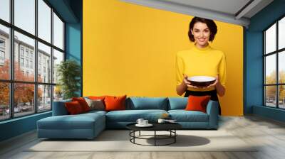 Young beautiful stylish woman holding an empty plate or dish isolated on yellow background with copy space Wall mural