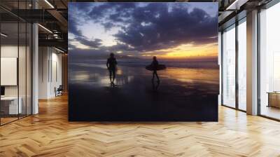 Silueta de dos amigos caminando con tabla de surf en la orilla de la playa con la última luz del atardecer en el horizonte en un día con sol, nubes y montañas con pueblo al fondo Wall mural