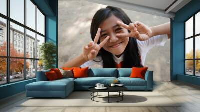 portrait of cute asian little girl with long black hair smiling with two fingers placed on face, or peace sign. Wall mural