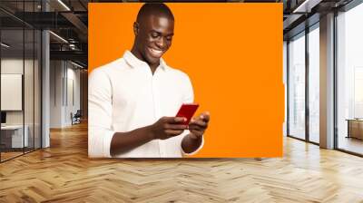 Generative AI : Happy overjoyed young african american man reading great news on smartphone amazed young guy receiving unexpected message on mobile phone isolated on orange studio background Wall mural