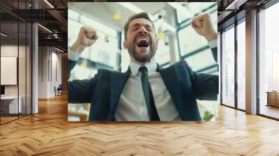 A man in a suit enthusiastically celebrates in a modern office, his fists raised in triumph, depicting professional accomplishment and joy. Wall mural