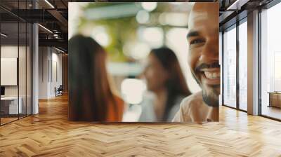 Shared laughter in soft focus, a man's smile is the foreground of an unfocused social gathering behind. Wall mural