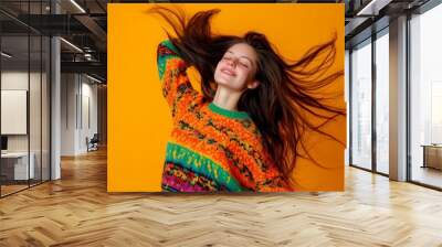 An excited young girl in an orange sweater jumps with fluttering hair in a studio. People's emotions and styles. Wall mural