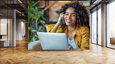 A cheerful Latina woman multitasks effortlessly, engaging in a phone conversation while diligently working on her laptop from the comfort of her home. Wall mural