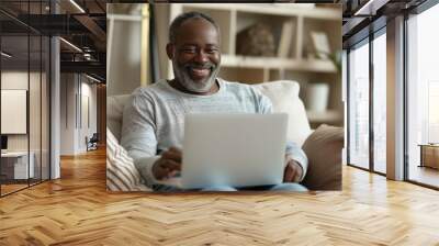 A happy African American middle-aged man is using a laptop while sitting on the sofa at home. The mature older user looks at the computer screen, browsing the internet and doing ecommerce shopping on Wall mural