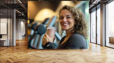 A middle-aged Brazilian woman smiling as she drinks a shake, surrounded by weights and cardio equipment in a well-equipped gym. Her positive energy reflects her healthy, balanced lifestyle. Wall mural