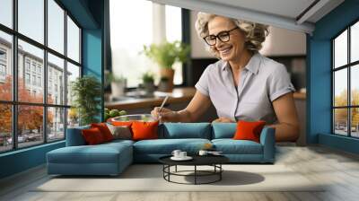 Smiling mature woman in eyeglasses cooking salad in kitchen at home Wall mural