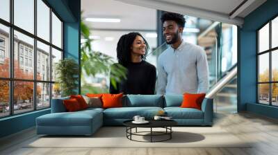 Two young Black colleagues, smiling and chatting, climb a modern office staircase. The background features a clean, well-lit office with stylish furniture and glass railings. Wall mural