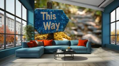 A close-up view of a rustic blue sign with the words 'This Way' on it, placed on a mossy ground, pointing towards a blurred, rocky path through a forest Wall mural