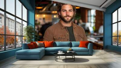 A confident young man with a beard and casual attire standing in a vibrant modern office environment with colleagues working in the background Wall mural