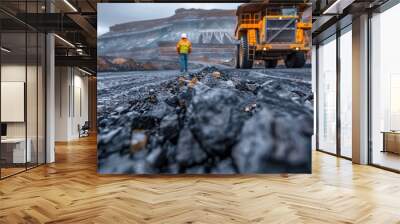 A person wearing safety gear is walking alongside a large industrial yellow mining truck in an open-pit mine with rocky, coal-laden terrain and layered geological formations Wall mural