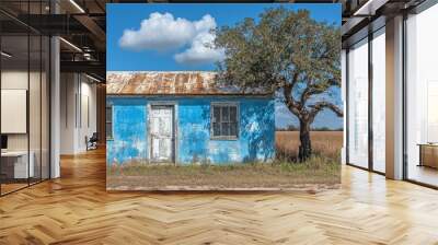 A vibrant blue rustic house stands under a large tree in a vast field, with a clear blue sky and fluffy clouds in the background, representing serene countryside life Wall mural