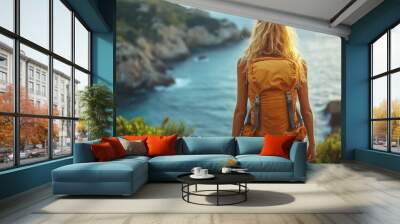 A woman with a bright orange backpack stands overlooking a cliffside view of the ocean, surrounded by lush greenery and rocky outcrops at sunset Wall mural