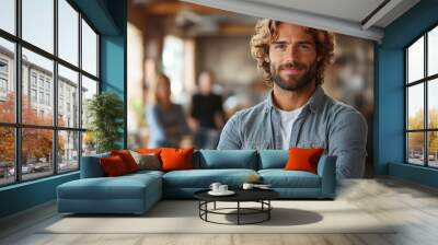 Portrait of a confident and approachable young man with curly blond hair standing with arms crossed in an open office environment with colleagues in the background Wall mural