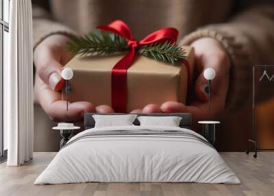 Person holding a beautifully wrapped Christmas gift with a red ribbon by a decorated tree Wall mural