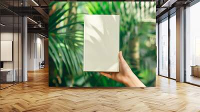 Woman hand holding empty white mockup of paperback book template on tropical trees background Wall mural