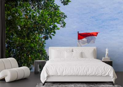 The red and white flag of Indonesia flutters among the trees against the blue sky Wall mural