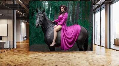 Portrait of young beautiful smiling brunette woman wearing pink silk dress riding dark horse at summer green forest. Wall mural