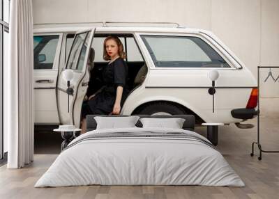 young beautiful girl in a black vintage polka dot dress next to a vintage white car with a vintage suitcase in her hands Wall mural