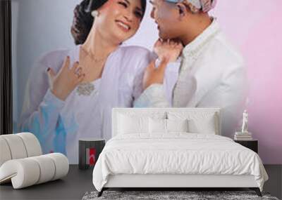 A pair of young Asian male and female lovers, standing smiling, woman showing affection to the man wearing modern Javanese traditional clothing, white velvet for the bride on a white studio background Wall mural