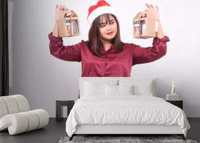 Beautiful young Southeast Asian woman smiling looking at box carrying 2 boxes of hamper gifts at Christmas wearing Santa hat modern red shirt outfit white background for promotion and advertising Wall mural