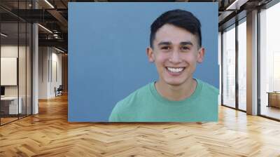 Close up portrait of a young hispanic teenager man looking at camera with a joyful smiling expression, against a blue background. Teenager being confident and smart. Wall mural