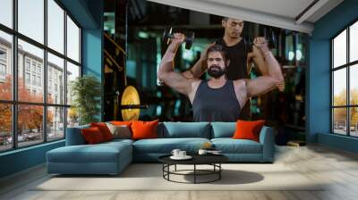 Caucasian man is using dumbbell in weight training for arm muscle inside gym with support from trainer to prevent injury dark background for exercising and workout concept Wall mural