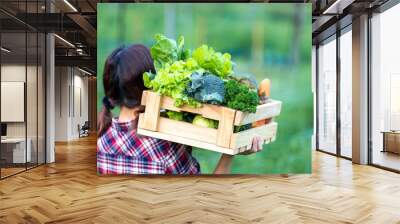 Woman farmer is carrying the wooden tray full of freshly pick organics vegetables over her head at the garden for harvest season and healthy diet food Wall mural