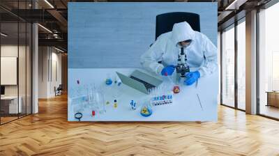 Asian medical scientist wearing protective white jumpsuit and face mask with glasses testing sample with microscope in clinical laboratory. Health care and medical concept. Virus pandemic concept.  Wall mural