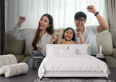 Asian family with father, mother and daughter sitting and watching TV and smile when watching fun program in TV in a comfortable couch in the livingroom at home. Family lifestyle relax  Wall mural