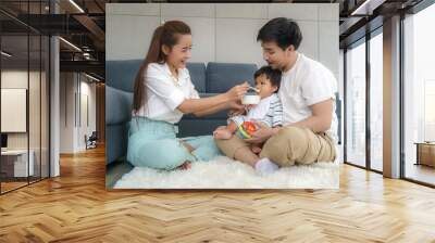 Asian mother feeding his 6 months old baby boy with solid food with spoon and father sitting near to cheer her son to eat food in living room at home.. Wall mural